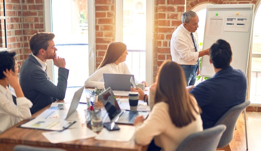 A man teaching something on a board to people sitting at a conference table