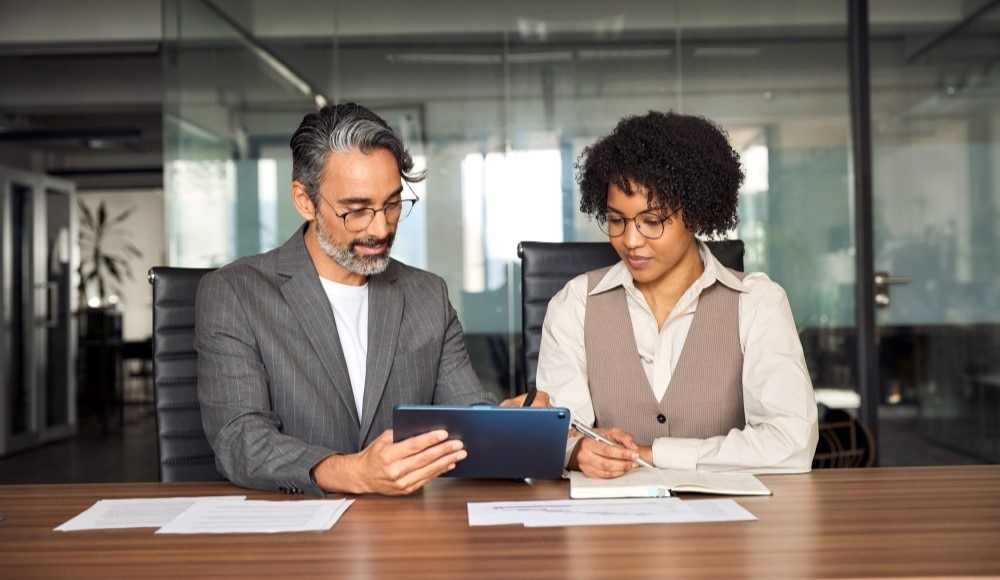 Trainer sitting at a conference table with a colleague and teaching her something on a tablet