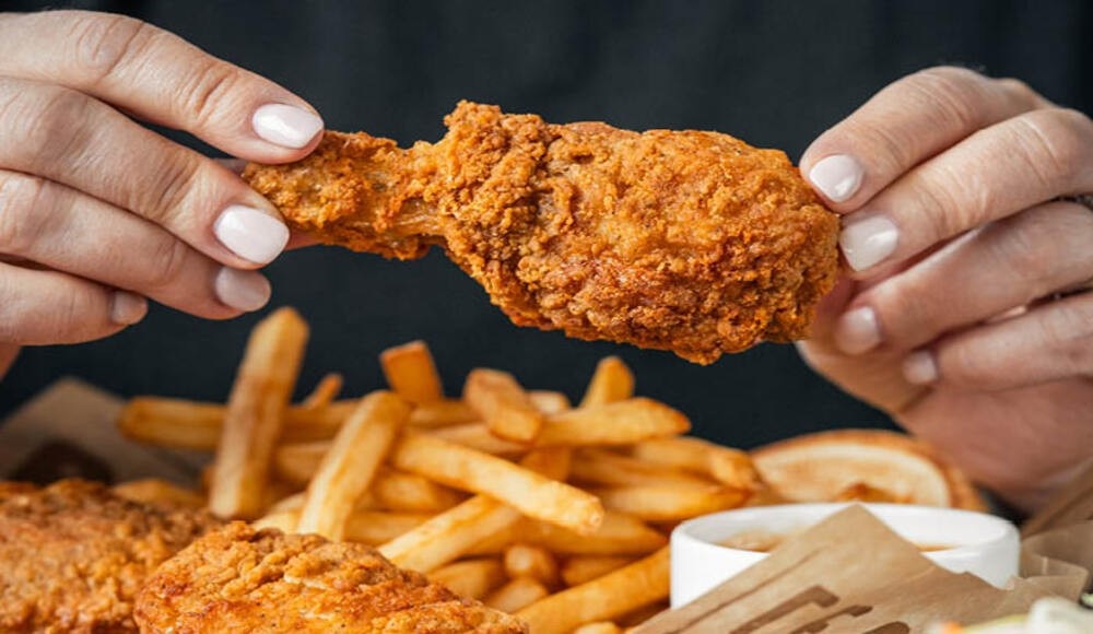 Visual of two hands holding a chicken drumstick above a plate full of fried chicken and fries.
