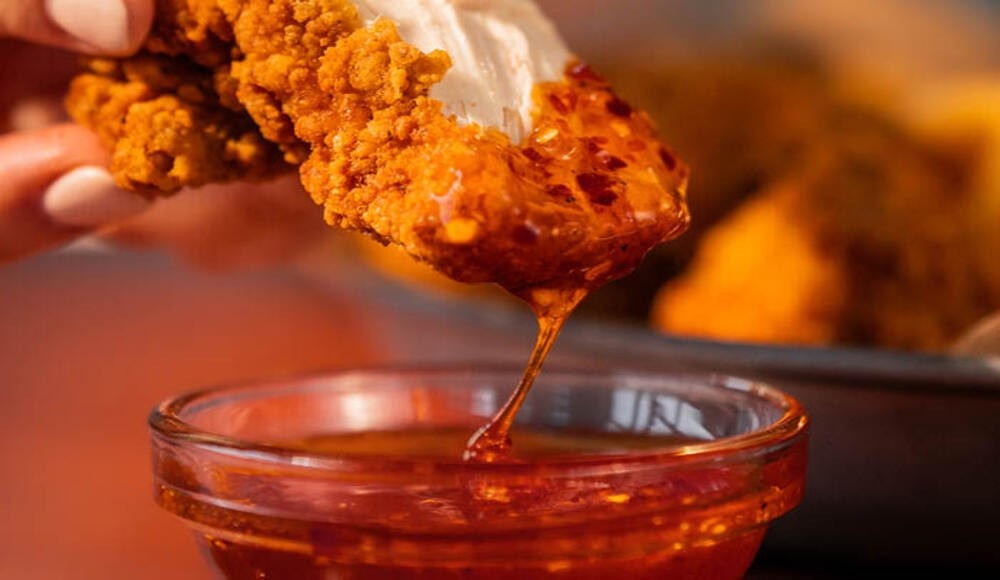 Visual of a piece of fried chicken being dipped in a ramekin full of honey piri-piri sauce.