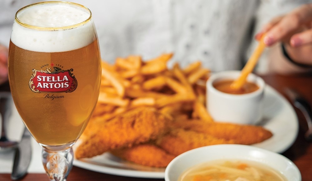 A man dips a fry in St-Hubert sauce. He is sitting at a table in front of a chicken breast strips meal and a chicken noodle soup. There is also a glass of Stella Artois.