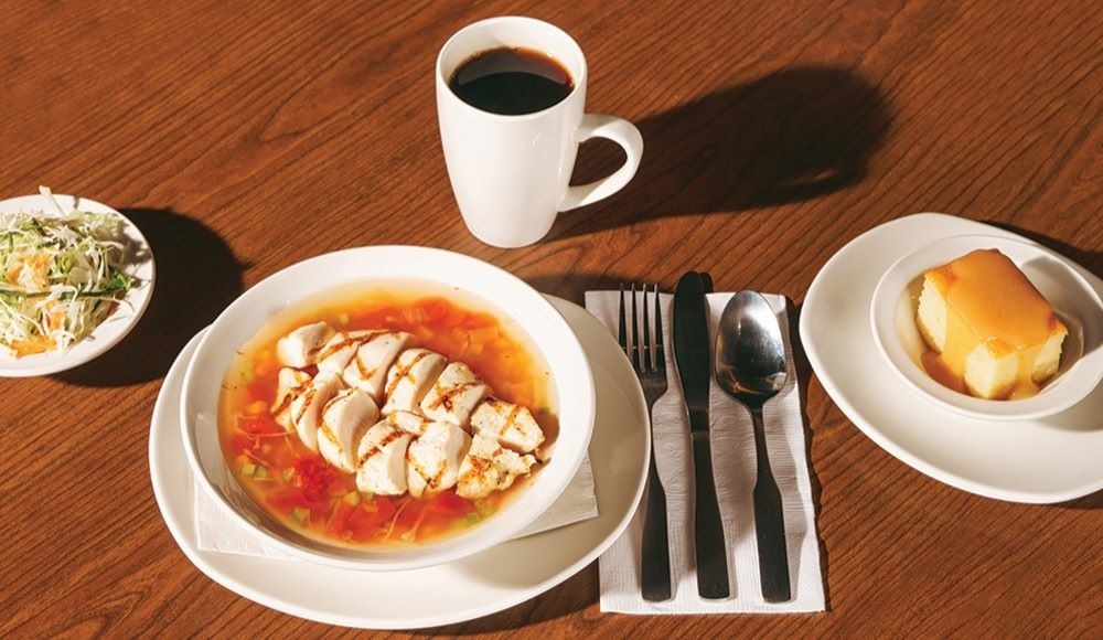 On a table, a full St-Hubert lunch with a hearty chicken and vegetable soup, coffee, traditional coleslaw and a vanilla cake.