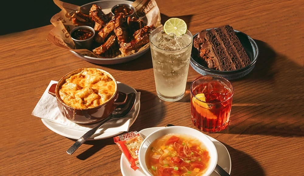 Onion soup, vegetable soup, crispy rib appetizer with a choice of sauces, decadent chocolate cake, ranch water and negroni - served on a wooden table.