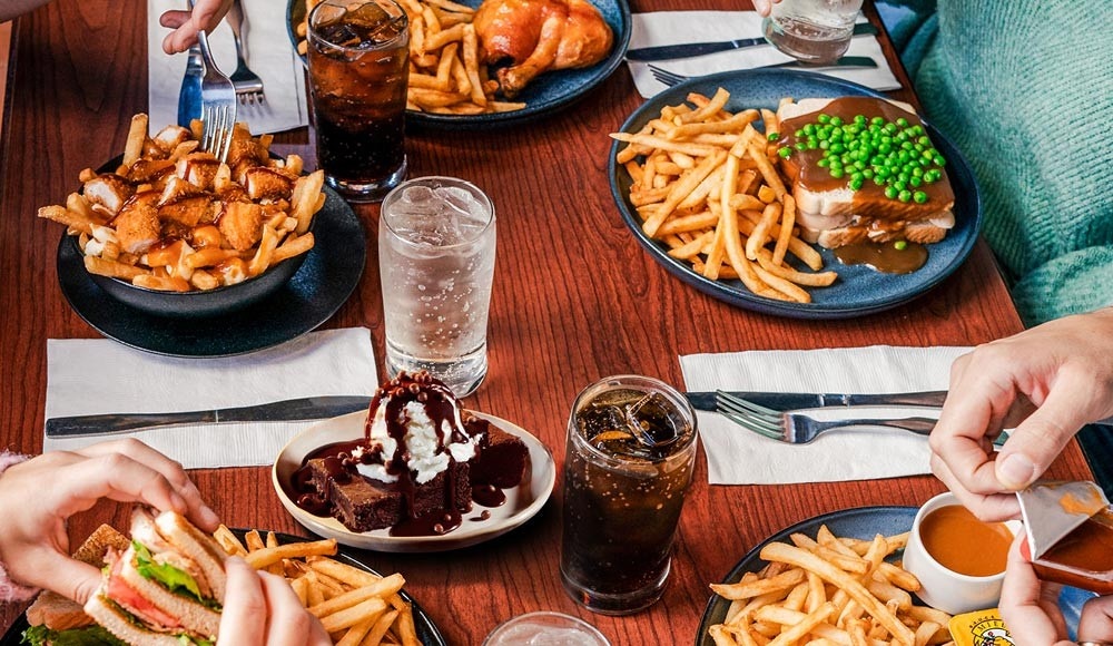 A fountain soft drink, a brownie, a St-Hubert poutine, a chicken leg, a club sandwich, and a hot chicken, all served on a wooden table